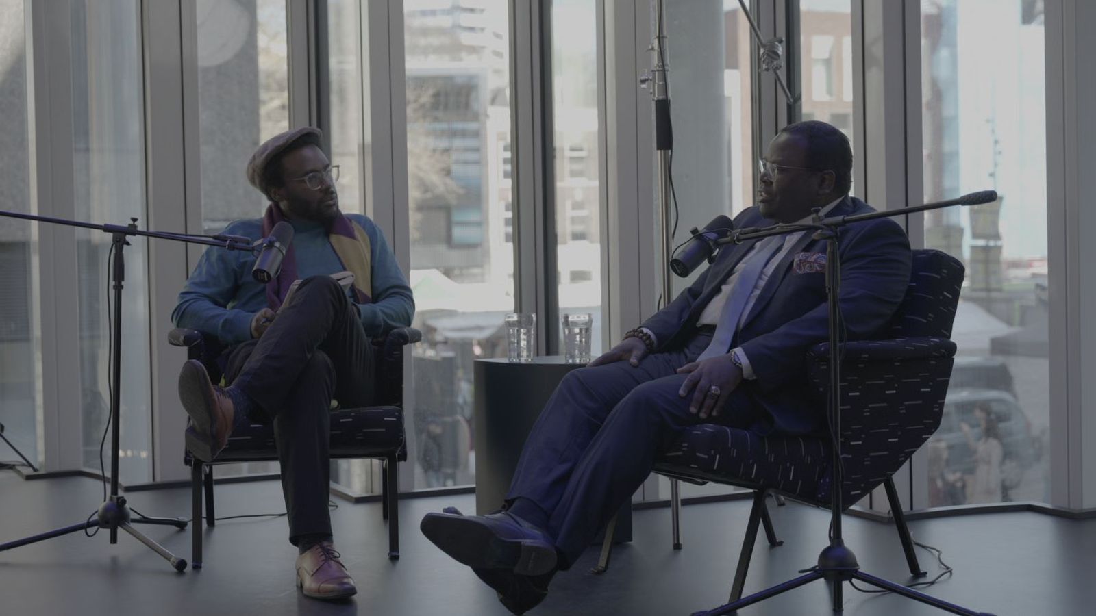 Presentator en gast in gesprek bij grote ramen, met microfoons en een ronde softbox